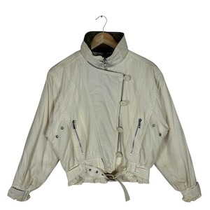Könnte beinhalten: Cremefarbene Vintage-Jacke mit hohem Kragen, Knopfverschluss und Reißverschlusstaschen. Die Jacke hat einen Gürtel in der Taille und gerippte Bündchen. Ein klassisches Oberbekleidungsstück.