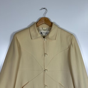 May include: A cream-coloured coat featuring a structured collar, long sleeves, and a front button closure with round, matching buttons. The garment displays distinct curved seam detailing across the chest and includes a Courr&egrave;ges Paris label.