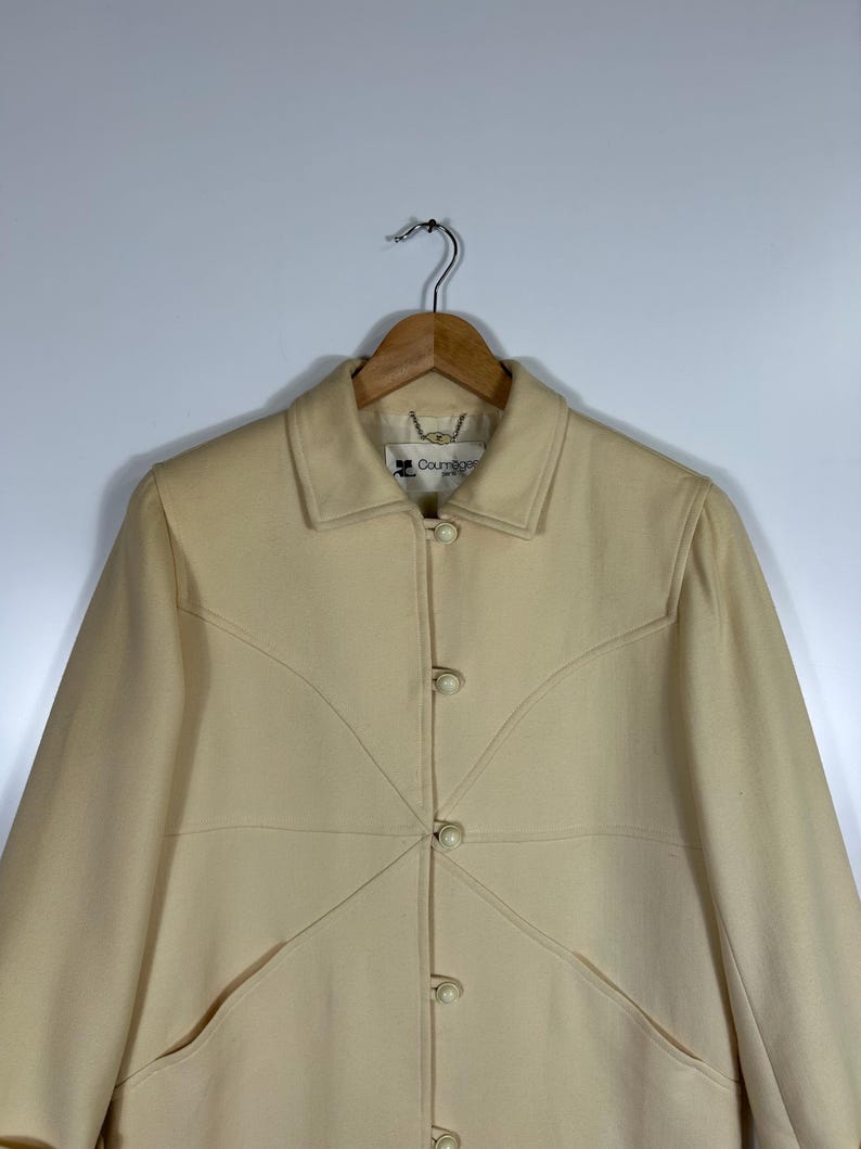 May include: A cream-coloured coat featuring a structured collar, long sleeves, and a front button closure with round, matching buttons. The garment displays distinct curved seam detailing across the chest and includes a Courr&egrave;ges Paris label.