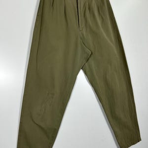 Puede incluir: Pantalones verde oliva con cierre de bot&oacute;n y cremallera, colgados de una percha de madera. Los pantalones tienen pliegues en la cintura y un &aacute;rea descolorida en la pierna izquierda. La tela parece ser ligera.