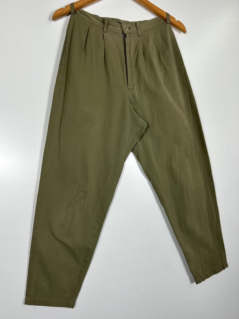 Puede incluir: Pantalones verde oliva con cierre de bot&oacute;n y cremallera, colgados de una percha de madera. Los pantalones tienen pliegues en la cintura y un &aacute;rea descolorida en la pierna izquierda. La tela parece ser ligera.