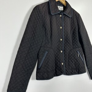 May include: A black quilted jacket with a diamond pattern, featuring a collar, button closure, and two front pockets with zippers. The jacket has navy blue trim and is hanging on a wooden hanger.
