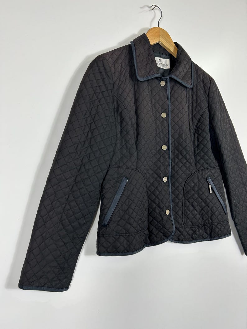May include: A black quilted jacket with a diamond pattern, featuring a collar, button closure, and two front pockets with zippers. The jacket has navy blue trim and is hanging on a wooden hanger.