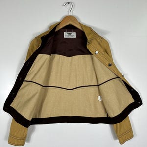 Peut inclure: Une veste beige avec une doublure int&eacute;rieure marron fonc&eacute; et une bordure marron fonc&eacute; le long des bords avant. La veste pr&eacute;sente un col, des manches longues et des boutons en m&eacute;tal argent&eacute;. Une &eacute;tiquette blanche avec le texte Courr&egrave;ges est visible au niveau du col.