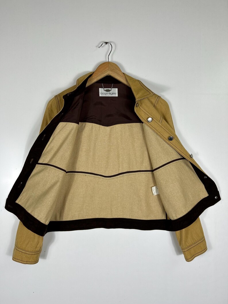 Peut inclure: Une veste beige avec une doublure int&eacute;rieure marron fonc&eacute; et une bordure marron fonc&eacute; le long des bords avant. La veste pr&eacute;sente un col, des manches longues et des boutons en m&eacute;tal argent&eacute;. Une &eacute;tiquette blanche avec le texte Courr&egrave;ges est visible au niveau du col.