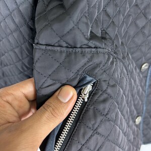 May include: Close-up of a dark gray quilted jacket with a silver zipper. The jacket has a diamond pattern and a blue trim. The zipper is open, revealing the interior lining. The jacket has silver buttons.