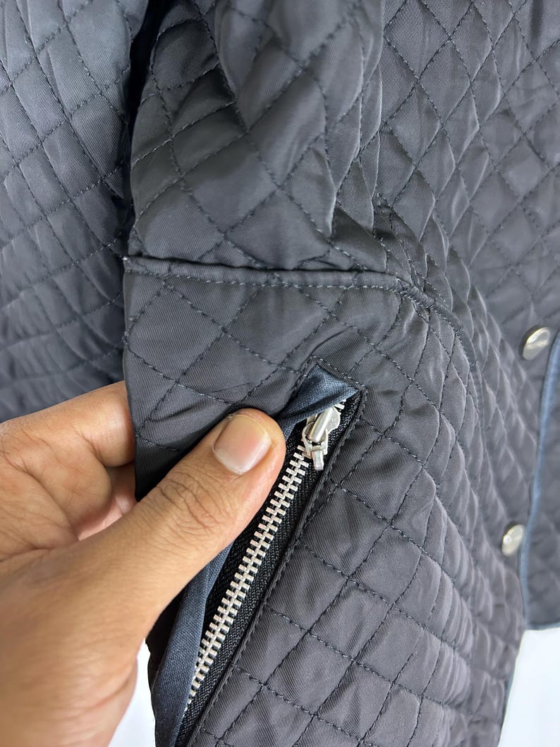 May include: Close-up of a dark gray quilted jacket with a silver zipper. The jacket has a diamond pattern and a blue trim. The zipper is open, revealing the interior lining. The jacket has silver buttons.