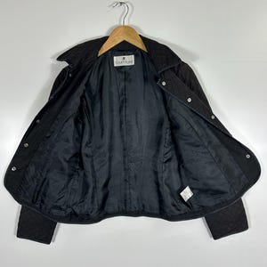 May include: Black quilted jacket with a collar and snap closures. The jacket is open, revealing a black lining and the brand label "courr&egrave;ges". The jacket is hanging on a wooden hanger against a white background.