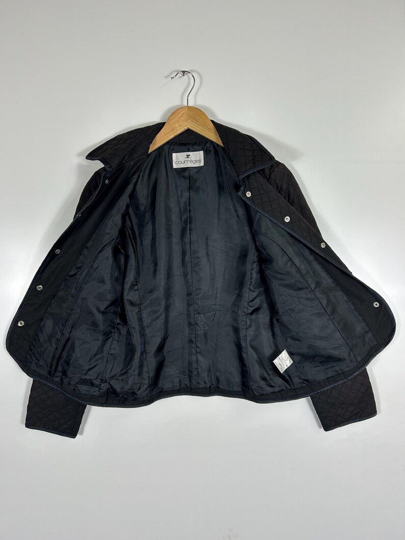 May include: Black quilted jacket with a collar and snap closures. The jacket is open, revealing a black lining and the brand label "courr&egrave;ges". The jacket is hanging on a wooden hanger against a white background.