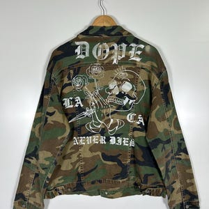 May include: Camouflage denim jacket with white graphic text and illustrations on the back. The design includes the word "DOPE", a skull, roses, and the phrase "LA CA NEVER DIES". The jacket is hanging on a wooden hanger.