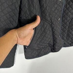 May include: Black quilted jacket with a diamond pattern. Features a zipper pocket and silver buttons. The jacket has a classic design and a dark color.