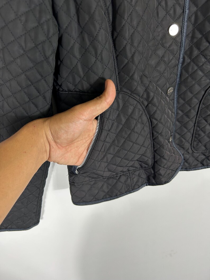May include: Black quilted jacket with a diamond pattern. Features a zipper pocket and silver buttons. The jacket has a classic design and a dark color.