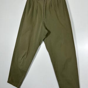 Puede incluir: Pantalones verde oliva con frente plisado, cierre de bot&oacute;n y corte relajado. Los pantalones cuelgan de una percha de madera sobre un fondo blanco. La tela parece ser ligera.