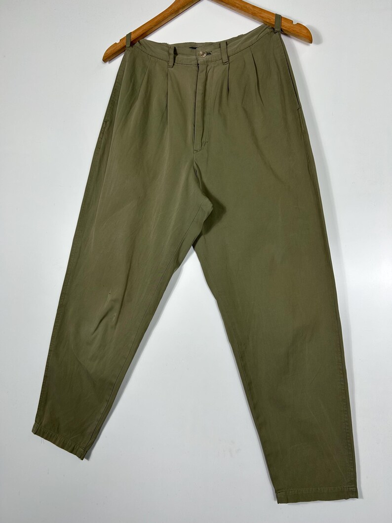 Puede incluir: Pantalones verde oliva con frente plisado, cierre de bot&oacute;n y corte relajado. Los pantalones cuelgan de una percha de madera sobre un fondo blanco. La tela parece ser ligera.