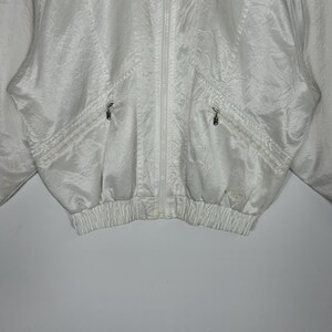 May include: A white, vintage-style jacket with a full zipper closure. The jacket features two front pockets with zippers and a ribbed hem. The fabric has a subtle sheen and a crinkled texture. The jacket is a light, neutral color.