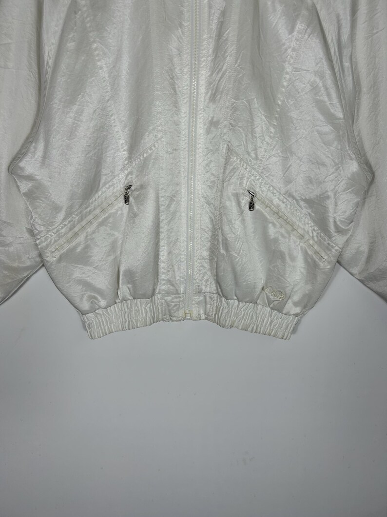 May include: A white, vintage-style jacket with a full zipper closure. The jacket features two front pockets with zippers and a ribbed hem. The fabric has a subtle sheen and a crinkled texture. The jacket is a light, neutral color.