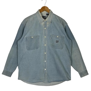 Puede incluir: Camisa vaquera azul claro con botones y mangas largas. Cuenta con dos bolsillos en el pecho y el logotipo "GAP" bordado en el bolsillo derecho. La camisa está colgada en una percha de madera.