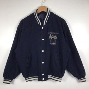 Puede incluir: Chaqueta universitaria azul marino con detalles blancos en el cuello, los puños y el dobladillo. La chaqueta tiene una parte delantera abotonada y un pequeño gráfico en el pecho con el texto "AND TIME TOGETHER".