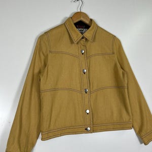 Peut inclure: Une veste &agrave; manches longues jaune moutarde dot&eacute;e d'un col pointu, d'une fermeture boutonn&eacute;e sur le devant avec des boutons en m&eacute;tal argent&eacute; et de surpiq&ucirc;res contrastantes marron fonc&eacute; le long des coutures, des poignets et de l'ourlet.