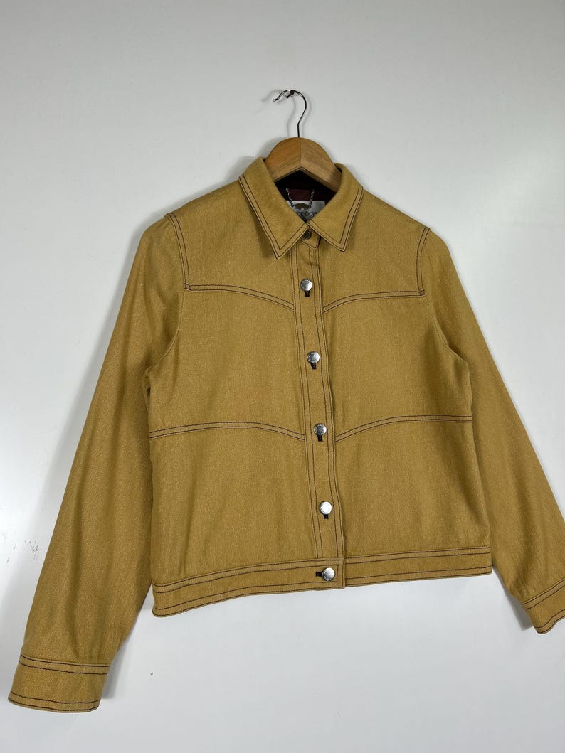 Peut inclure: Une veste &agrave; manches longues jaune moutarde dot&eacute;e d'un col pointu, d'une fermeture boutonn&eacute;e sur le devant avec des boutons en m&eacute;tal argent&eacute; et de surpiq&ucirc;res contrastantes marron fonc&eacute; le long des coutures, des poignets et de l'ourlet.
