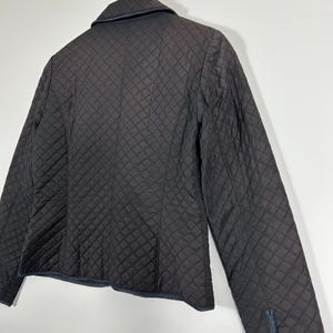May include: A black quilted jacket with a classic design. The jacket features a diamond pattern throughout the fabric, a collar, and long sleeves. The jacket is hanging on a wooden hanger against a white background.