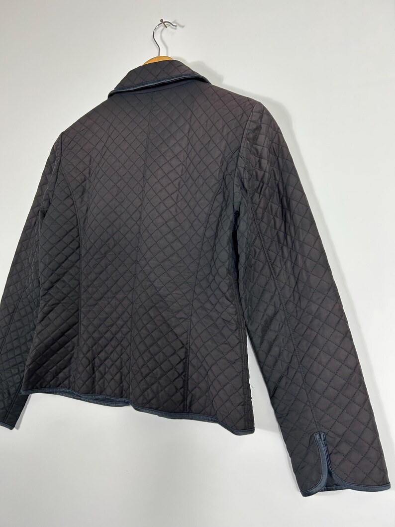 May include: A black quilted jacket with a classic design. The jacket features a diamond pattern throughout the fabric, a collar, and long sleeves. The jacket is hanging on a wooden hanger against a white background.