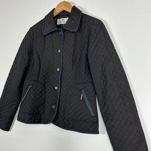 May include: Black quilted jacket with a diamond pattern, featuring a collar, button closure, and two front pockets. The jacket has navy blue trim and a small zipper pocket. The jacket is hanging on a wooden hanger.