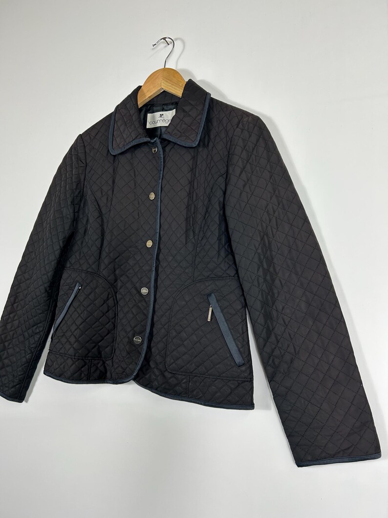 May include: Black quilted jacket with a diamond pattern, featuring a collar, button closure, and two front pockets. The jacket has navy blue trim and a small zipper pocket. The jacket is hanging on a wooden hanger.