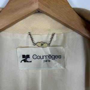 May include: The interior of a beige garment featuring a white fabric label with the black text Courr&egrave;ges paris and a logo. A silver-toned metal chain hanger loop is attached above the label, featuring a small decorative tag with the same logo.