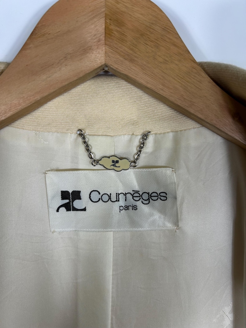 May include: The interior of a beige garment featuring a white fabric label with the black text Courr&egrave;ges paris and a logo. A silver-toned metal chain hanger loop is attached above the label, featuring a small decorative tag with the same logo.