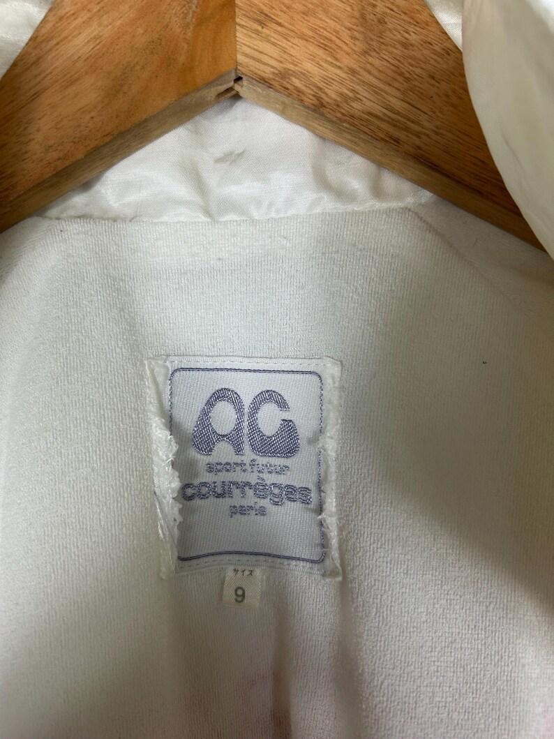May include: Close-up of a white jacket with a label that reads "AC sport futur courr&egrave;ges paris" in a light blue font. The label also has the number "9" printed on it. The jacket is hanging on a wooden hanger.