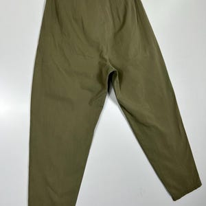 Puede incluir: Pantalones verde oliva colgados de una percha de madera. Los pantalones tienen cintura alta, trabillas y un corte relajado. La tela parece ser un material ligero, adecuado para climas c&aacute;lidos. Los pantalones se muestran sobre un fondo blanco.