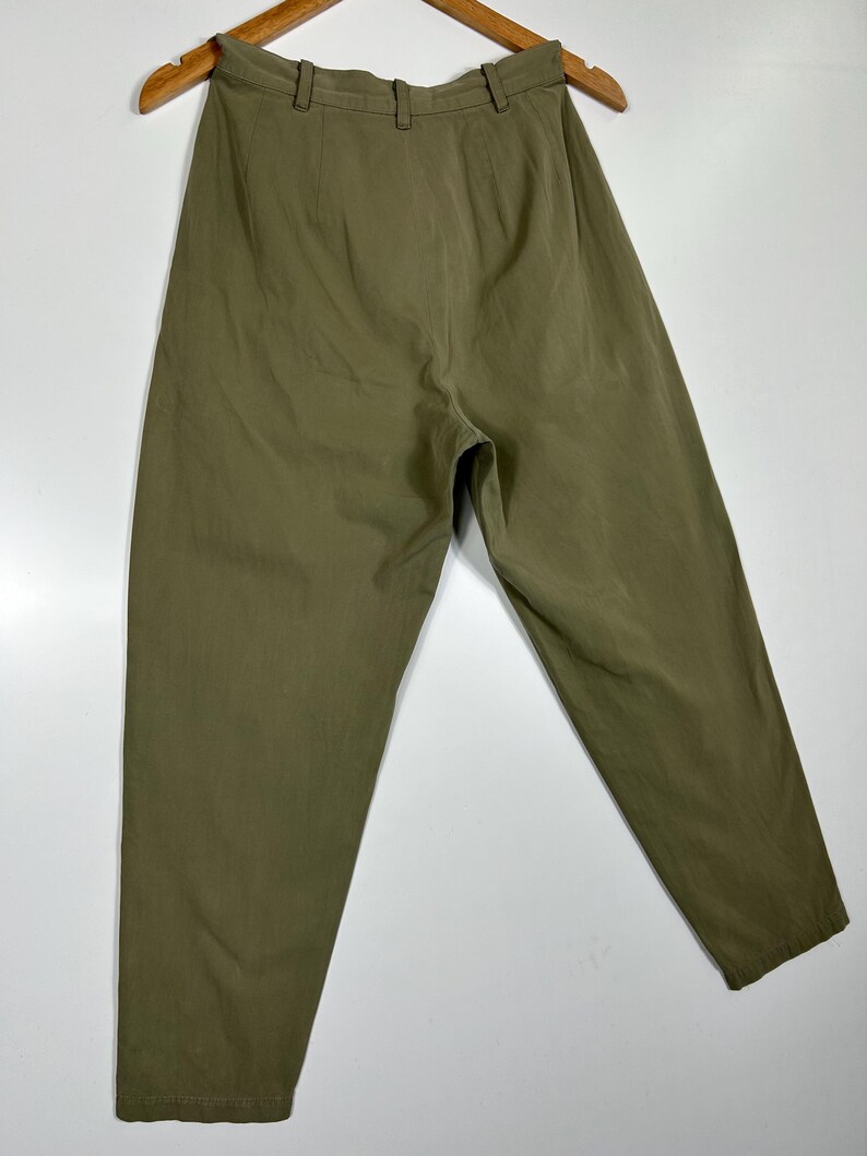 Puede incluir: Pantalones verde oliva colgados de una percha de madera. Los pantalones tienen cintura alta, trabillas y un corte relajado. La tela parece ser un material ligero, adecuado para climas c&aacute;lidos. Los pantalones se muestran sobre un fondo blanco.