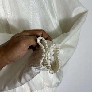 May include: Close-up of a white jacket sleeve. The jacket is made of a shiny, lightweight material. The sleeve has an elastic cuff and a gathered hem. The jacket appears to be new or in excellent condition.