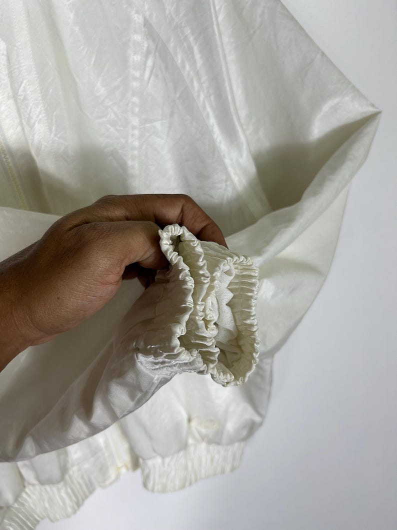 May include: Close-up of a white jacket sleeve. The jacket is made of a shiny, lightweight material. The sleeve has an elastic cuff and a gathered hem. The jacket appears to be new or in excellent condition.