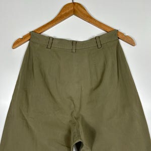 Puede incluir: Pantalones verde oliva colgados de una percha de madera. Los pantalones tienen cintura alta y dise&ntilde;o de pernera ancha. La tela parece ser un material ligero, adecuado para climas c&aacute;lidos. Los pantalones se muestran sobre un fondo blanco liso.