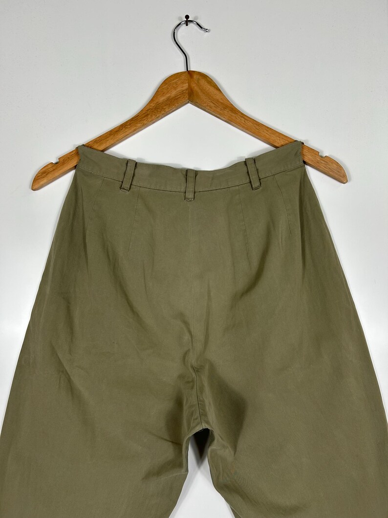Puede incluir: Pantalones verde oliva colgados de una percha de madera. Los pantalones tienen cintura alta y dise&ntilde;o de pernera ancha. La tela parece ser un material ligero, adecuado para climas c&aacute;lidos. Los pantalones se muestran sobre un fondo blanco liso.