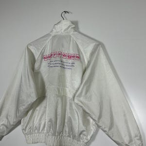 May include: White nylon jacket with a high collar and a full-length zipper. The back of the jacket features the word "carr&egrave;ges" in pink block letters, along with additional text. The jacket has a ribbed hem and cuffs.