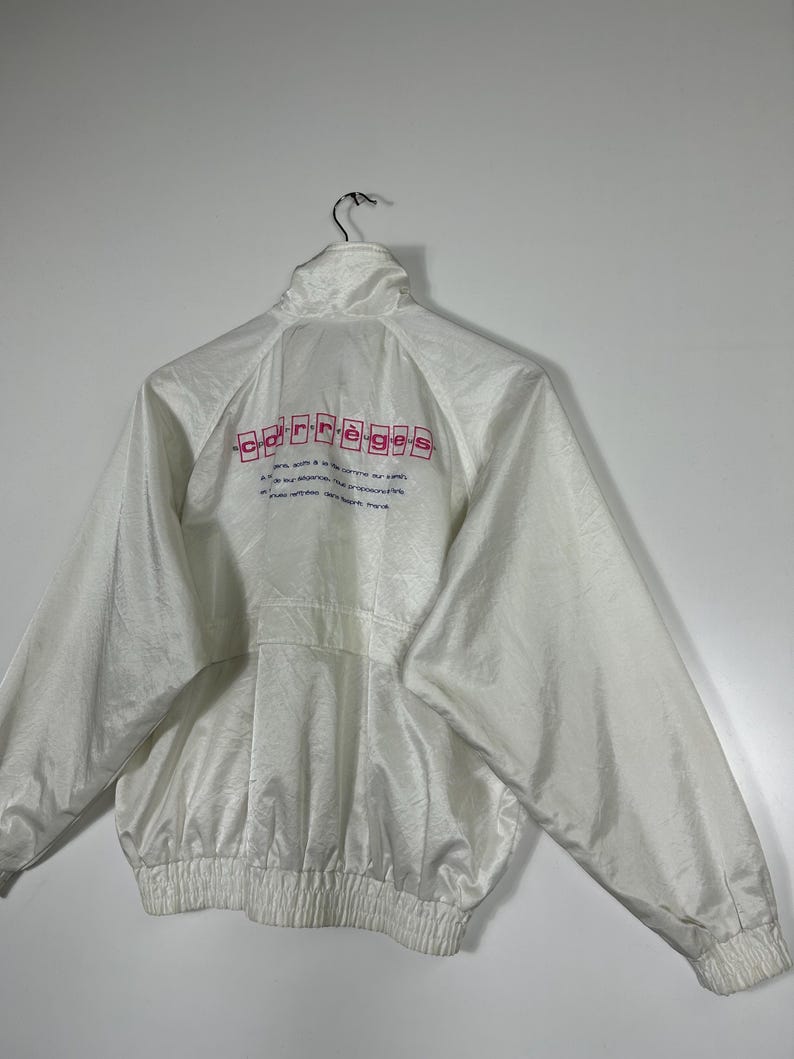 May include: White nylon jacket with a high collar and a full-length zipper. The back of the jacket features the word "carr&egrave;ges" in pink block letters, along with additional text. The jacket has a ribbed hem and cuffs.