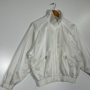 May include: A white vintage track jacket with a high collar and a full-length zipper. The jacket has two front pockets with zippers and ribbed cuffs and hem. The fabric has a subtle sheen. The jacket is hanging on a wooden hanger.