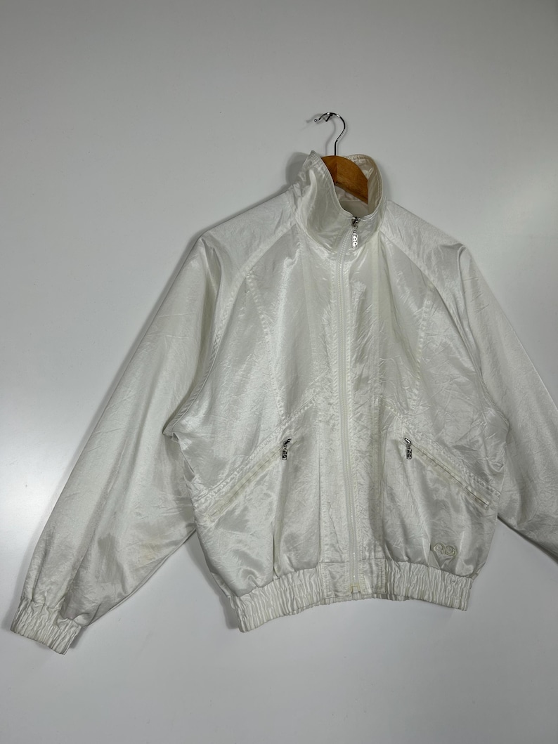 May include: A white vintage track jacket with a high collar and a full-length zipper. The jacket has two front pockets with zippers and ribbed cuffs and hem. The fabric has a subtle sheen. The jacket is hanging on a wooden hanger.