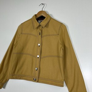 Peut inclure: Une veste &agrave; manches longues jaune moutarde dot&eacute;e d'un col pointu, d'une fermeture boutonn&eacute;e sur le devant avec des boutons m&eacute;talliques argent&eacute;s et de surpiq&ucirc;res contrast&eacute;es marron fonc&eacute; le long des coutures, des panneaux de poitrine et des poignets.