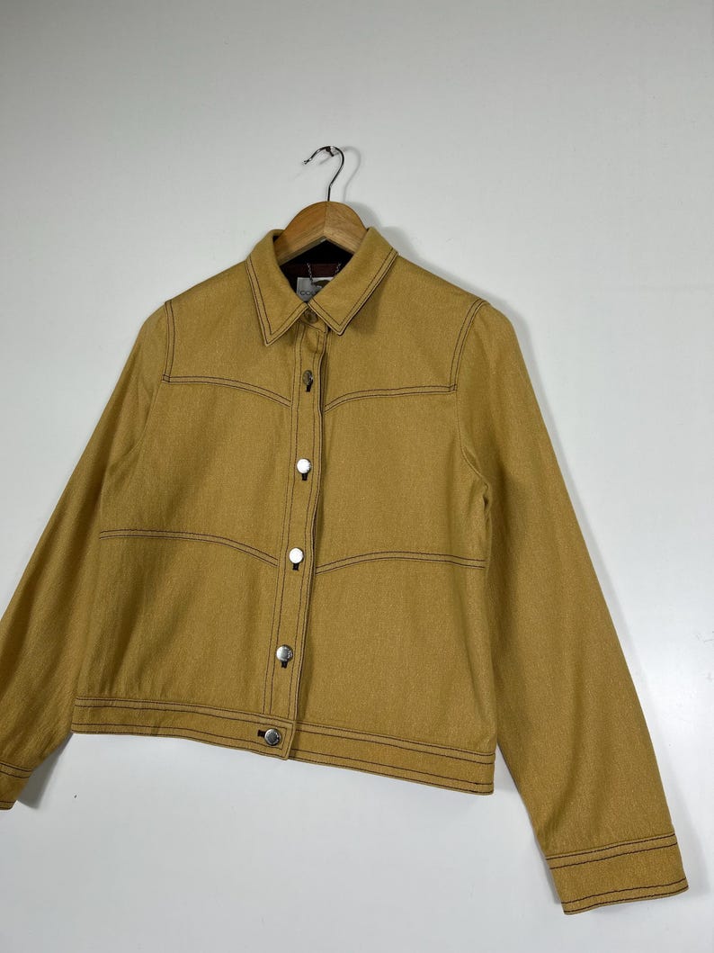 Peut inclure: Une veste &agrave; manches longues jaune moutarde dot&eacute;e d'un col pointu, d'une fermeture boutonn&eacute;e sur le devant avec des boutons m&eacute;talliques argent&eacute;s et de surpiq&ucirc;res contrast&eacute;es marron fonc&eacute; le long des coutures, des panneaux de poitrine et des poignets.