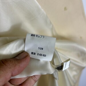 May include: A cream-coloured garment interior featuring a white fabric tag with the text "裏地 キュプラ", "SUN", and "製造 C-OS-168". The tag is attached to a smooth, satin-like lining material, providing clear details on the garment's internal construction and labelling.