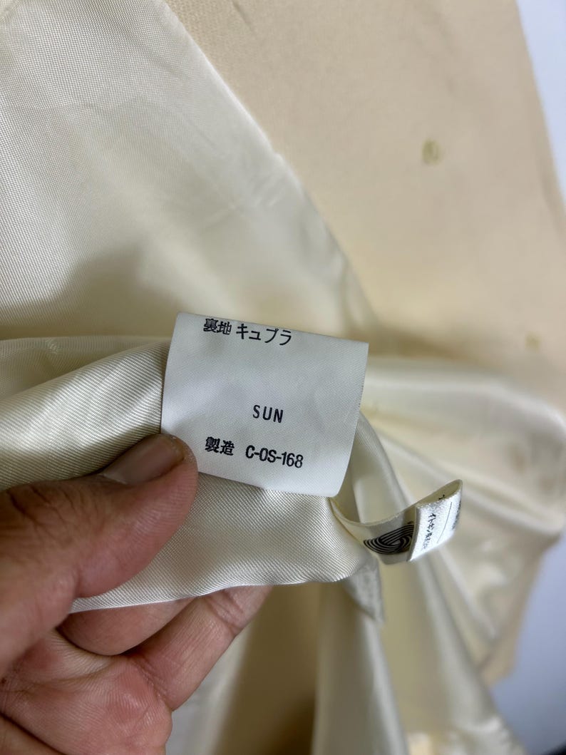 May include: A cream-coloured garment interior featuring a white fabric tag with the text "裏地 キュプラ", "SUN", and "製造 C-OS-168". The tag is attached to a smooth, satin-like lining material, providing clear details on the garment's internal construction and labelling.