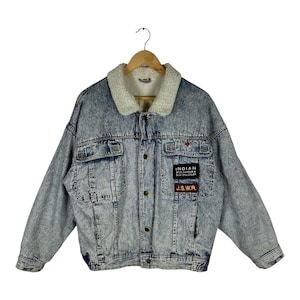 May include: A light blue acid-wash denim jacket featuring a white sherpa collar, long sleeves, and a button-up front. The chest displays two rectangular patches with the text "INDIAN RIDE HARDER & STAY ON LONGER" and "J.S.W.R." in white and red.