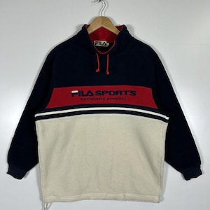 May include: A navy blue, red, and cream-colored FILA SPORTS fleece pullover. The long-sleeved top features a red band across the chest with white text and a white stripe. The collar is red with a navy blue exterior.