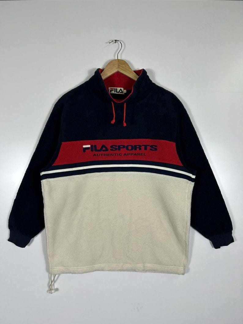 May include: A navy blue, red, and cream-colored FILA SPORTS fleece pullover. The long-sleeved top features a red band across the chest with white text and a white stripe. The collar is red with a navy blue exterior.