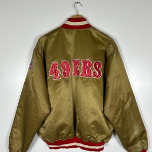 May include: A gold satin bomber jacket with red and white trim. The back of the jacket features the embroidered text "49ERS" in red. The jacket has a ribbed collar, cuffs, and hem with red and white stripes. A small patch is visible on the left sleeve.