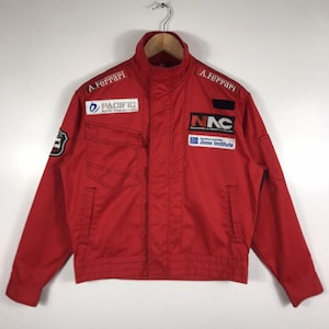 以下が含まれることがあります： 「A. Ferrari」、「PACIFIC RACING TEAM」、「NAC NARANJOS AUTOMOTIVE COLLEGE」などのパッチとテキストが特徴の赤いレーシングジャケット。 ジャケットはハイカラー、2つのフロントポケット、ジッパー開閉式です。 木製ハンガーにかかっています。